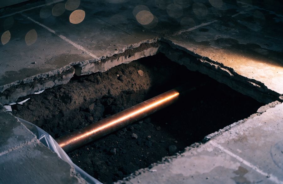 Section of concrete floor cut away to expose damp soil and a copper water line running below slab level