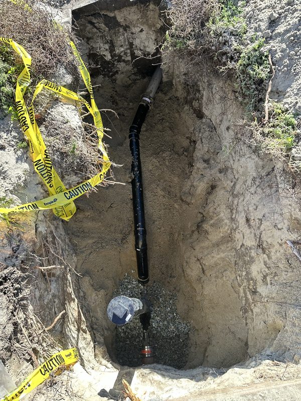 Open excavation with a new black sewer line and a two-way cleanout bedded in gravel, joined with banded couplings, caution tape across the edge of the hole