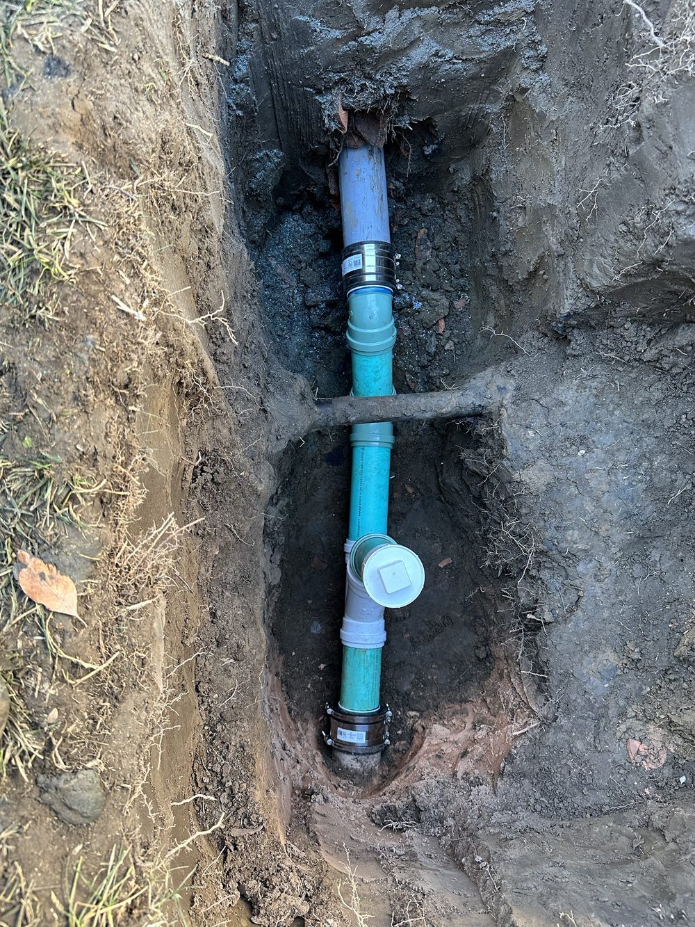 Trench showing new green PVC sewer pipe with a white two-way cleanout, connected at both ends to the existing pipe with banded rubber couplings