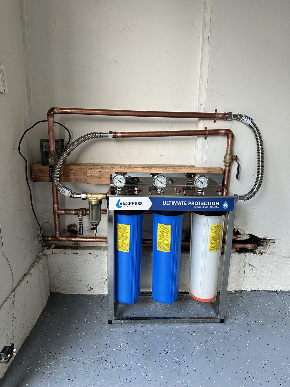 Three-stage whole house water filtration system mounted on a plywood backer with copper supply piping, pressure gauges, isolation valves and a pressure regulator