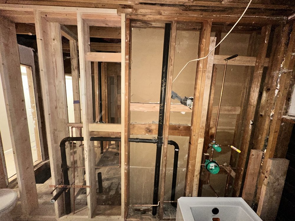 Bathroom rough in with open stud walls showing black ABS drain and vent piping, copper water lines and a shower valve with protective caps, next to a new shower pan