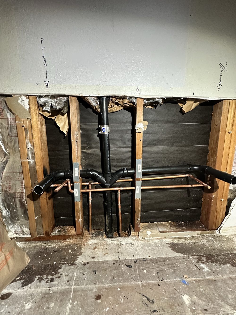Section of wall opened to expose a new black ABS drain, waste and vent assembly with copper water lines, strapped to the studs with metal protection plates