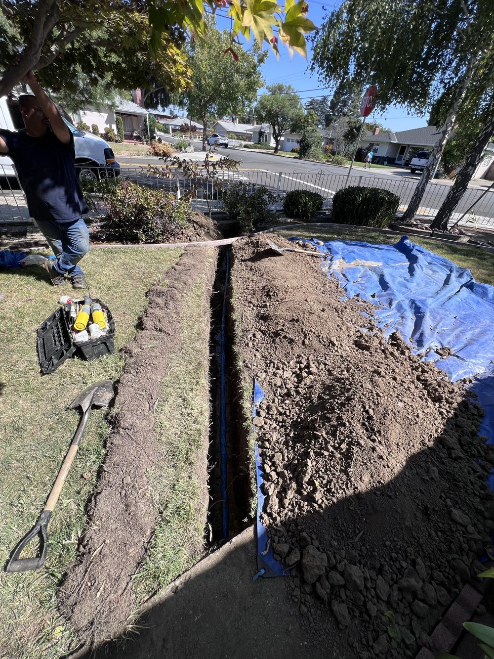 Narrow trench cut across a front lawn for a water service line, excavated soil stacked on blue tarps to protect the grass, with a plumber and hand tools beside it