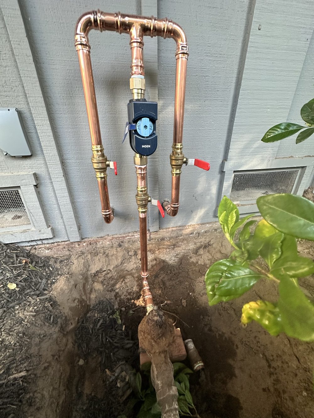 New copper water service manifold with three brass ball valves and a Moen Flo smart shutoff, mounted on a house exterior above an open excavation where it ties into the old galvanized line